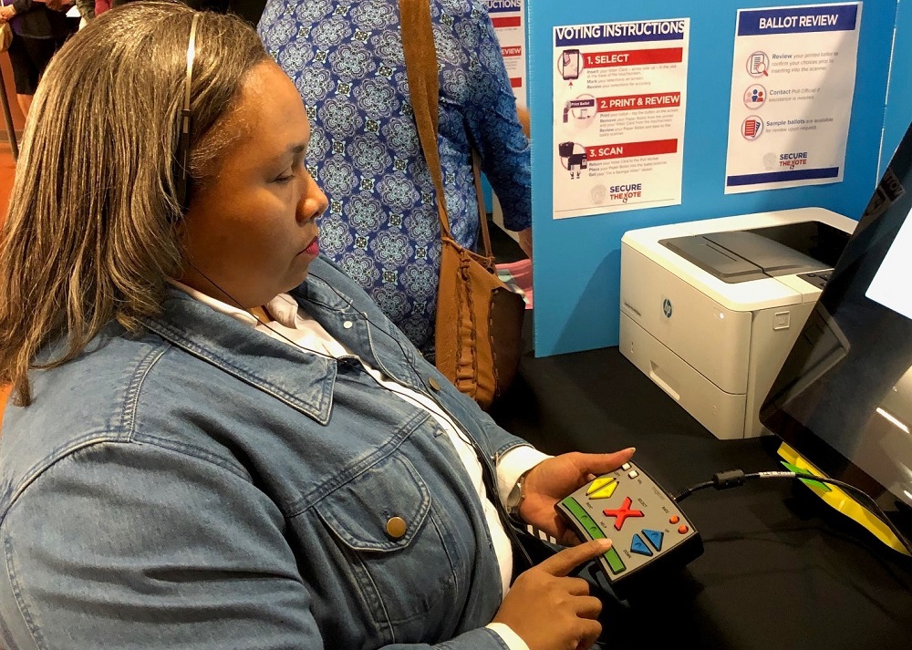 Empish at voting table,  wearing headset and holding key pad for voting machine in left hand, using right index finger to operate the key pad pushing the buttons. This picture was taken before the COVID-19 epidemic and she is not wearing a mask.