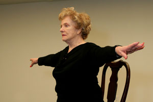 older woman standing with arms outstretched with chair behind her