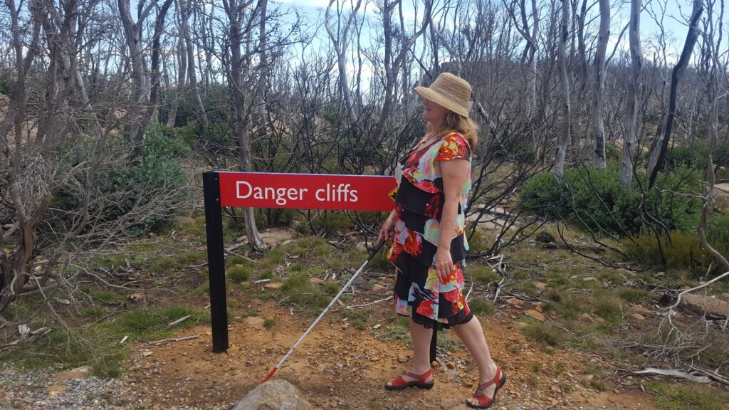 maribel standing on cliff next to sign "Danger cliffs". She is wearing a hat and holding out her white cane. photo by Harry Williamson