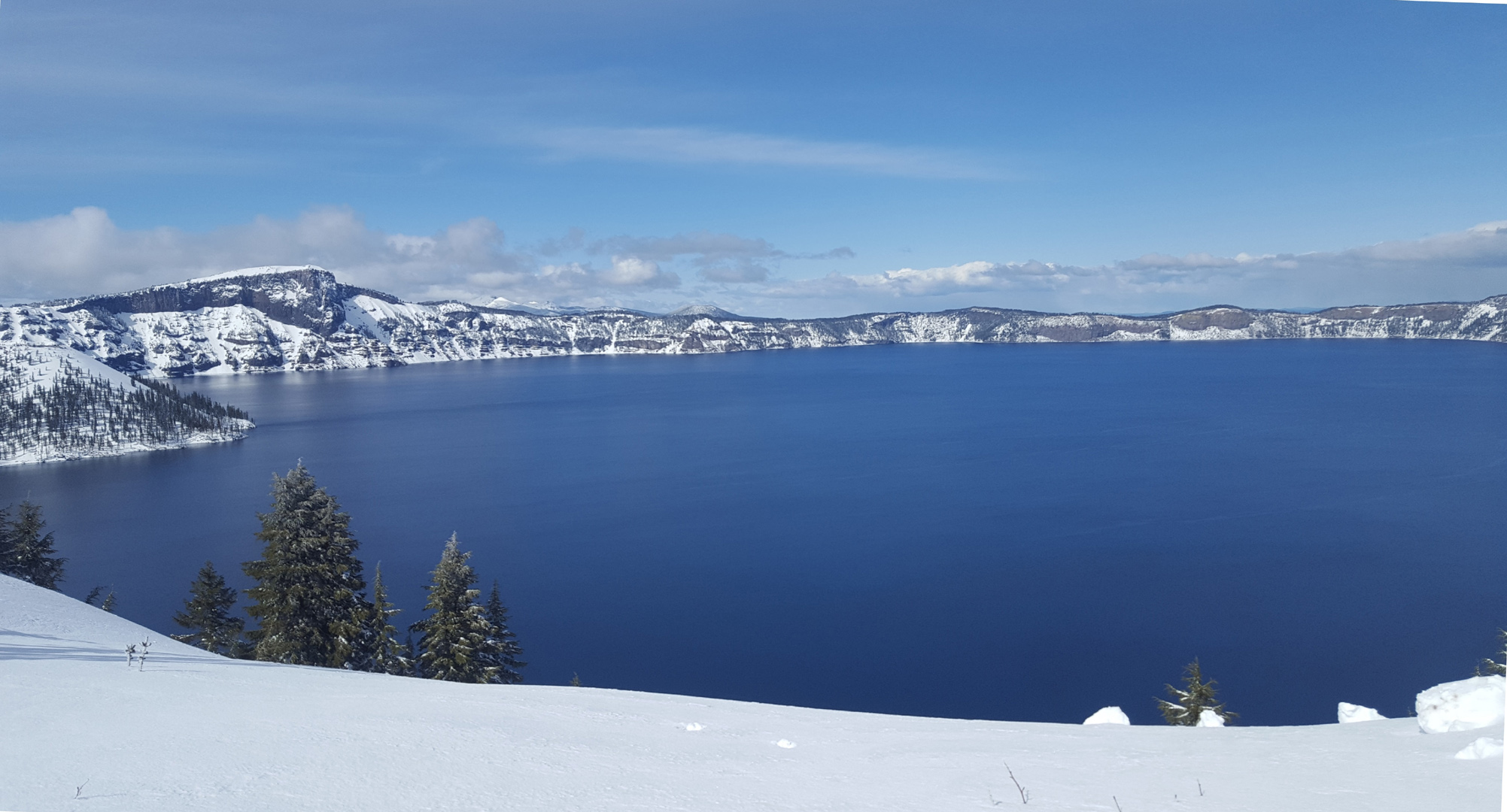 picture of Crater Lake, a very blue lake surrounded by snow covered  shore, mountains and clouds in the back ground. Photo by Harry Williamson