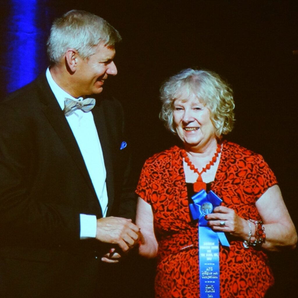 Lynda with Craig Meador at APH winning award for artwork. She is holding a blue ribbon and wearing a red dress and red necklace.