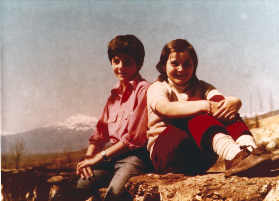 Maribel and brother as children seated next to each other on mountaintop with snow covered mountain in background
