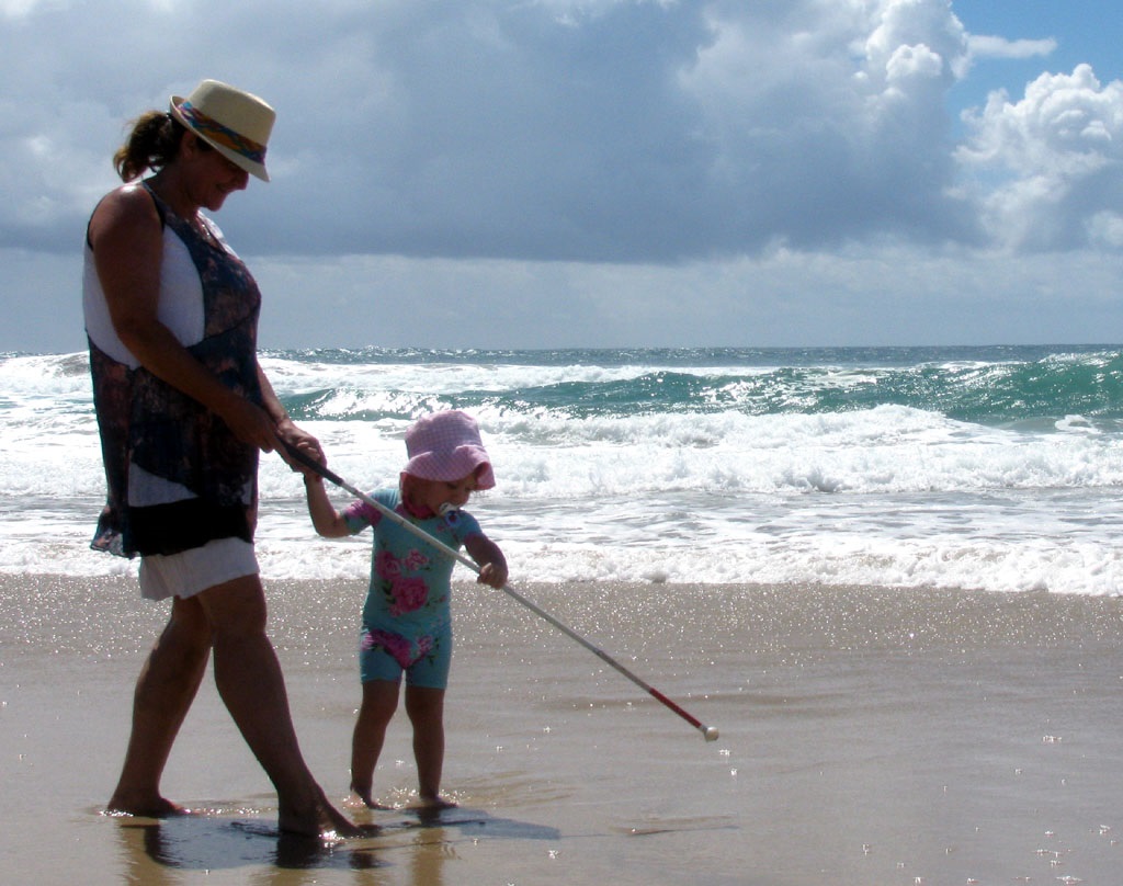 Maribel and child walking on a beach. Maribel is holding a long white cane and the child is trying to direct the cane in front of Maribel.