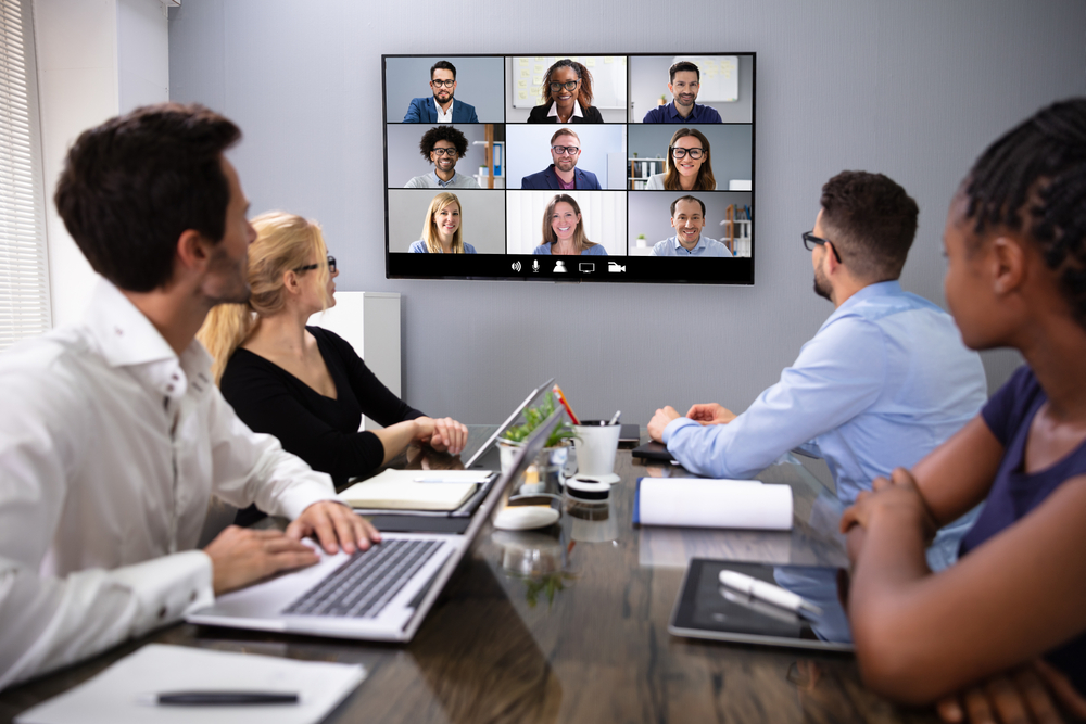 group of people with computers watching a tv screen with others in a zoom type of meeting.
